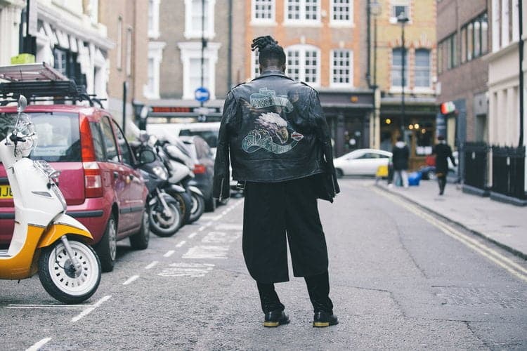 Un créateur de mode marche avec assurance dans la rue, vêtu d'une veste en cuir, montrant son timing impeccable et son sens de la mode.