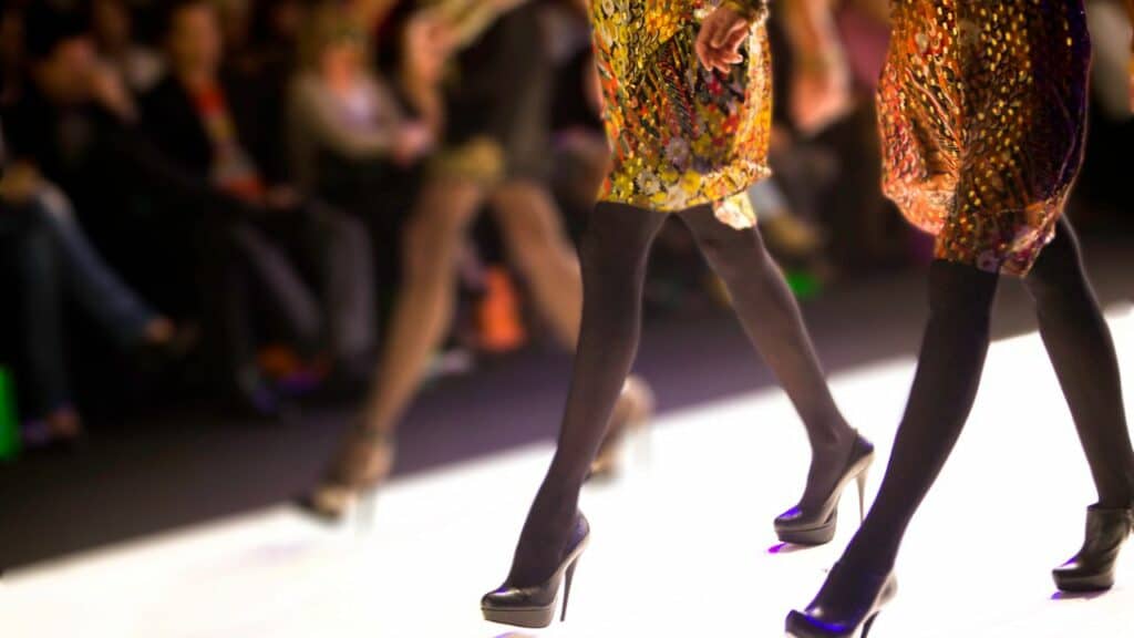 Two strutting women along a catwalk in high heels during a fashion show featuring resort cruises and pre-collection designs.