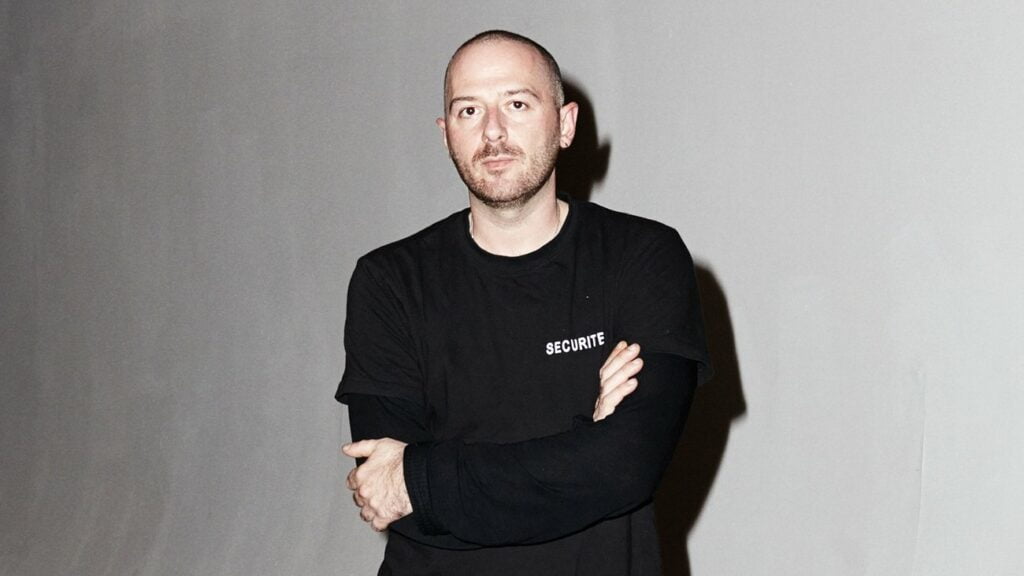 Ein glatzköpfiger Mann in einem schwarzen Hemd steht vor einer weißen Wand und repräsentiert das millenniale Verständnis des Designers Demna Gvasalia.