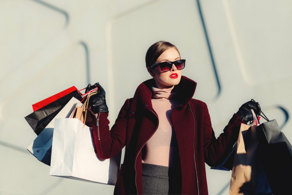 Une femme présente des sacs de courses devant un bâtiment, soulignant la fusion des marques de mode à l'ere numérique.