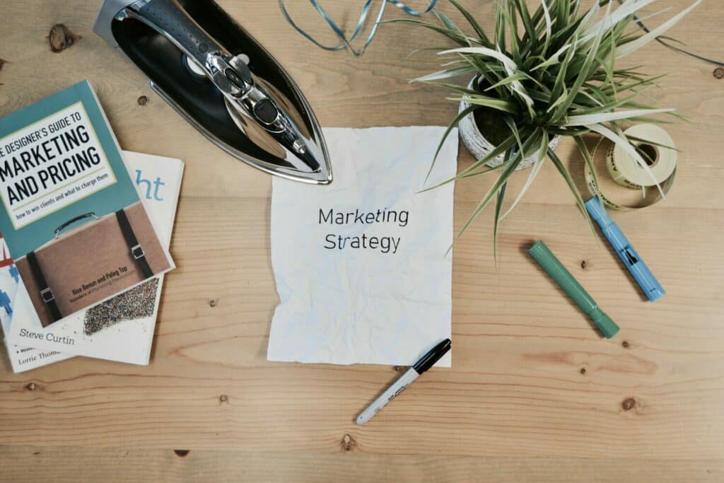 Sobre una mesa de madera hay una nota con la palabra "estrategia de marketing" en la que se destacan los 4 pasos fundamentales para tener éxito como aspirante a diseñador.