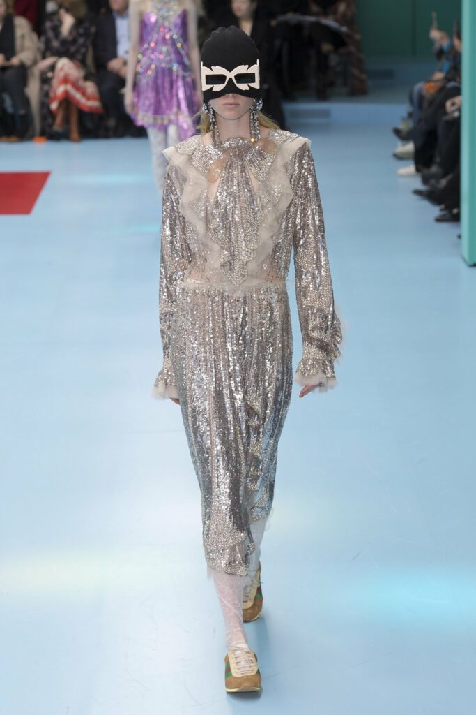 A woman in a sequin dress parades down the AW 2018 catwalk, embodying an 80s party aesthetic.