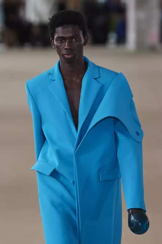 Ein Mann in einem blauen Mantel zeigt auf dem Laufsteg die ikonischen Modetrends des Jahres 2023.
