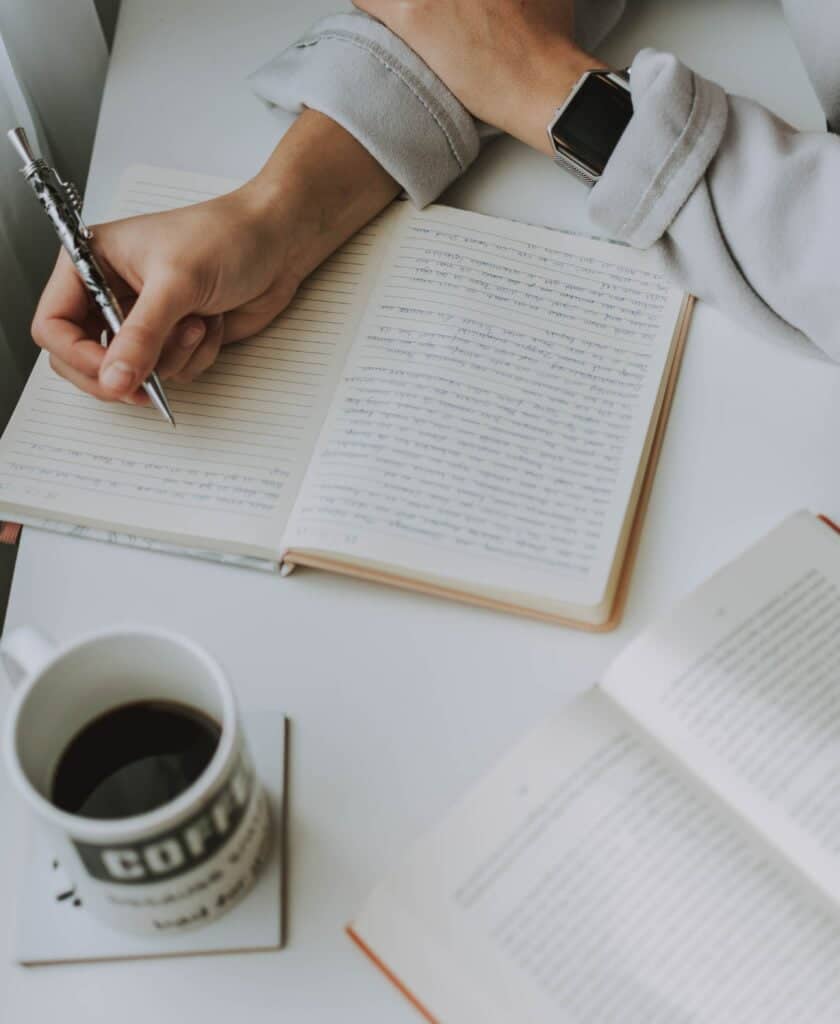 Una mujer escribe en un cuaderno con una taza de café mientras prepara un dossier de prensa.