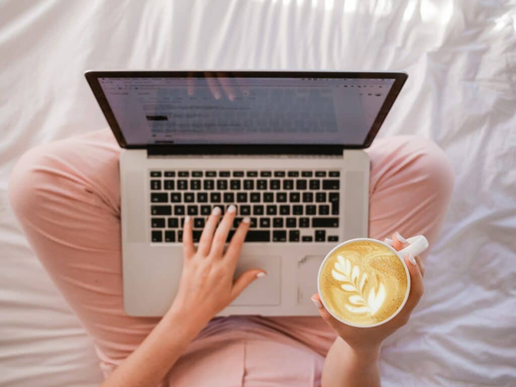 A woman sits on a bed with a laptop, embodying the role of fashion blogger and content creator.