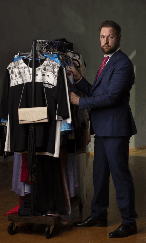 一个穿着西装、打着领带的男人站在衣帽架旁，在一间专为时尚而设的办公室里踌躇满志，立志成为一名时装设计师。