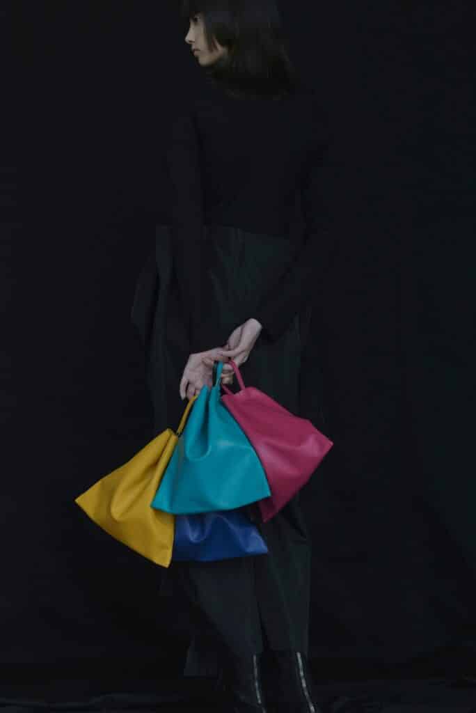 A woman elegantly shows off fashion trends 2024 with several colourful bags on an elegant black background.