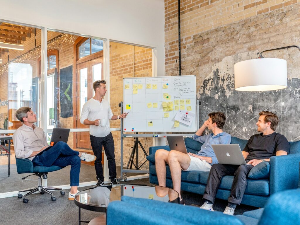 drei Männer sitzen an Laptops und beobachten den Mann neben dem Whiteboard