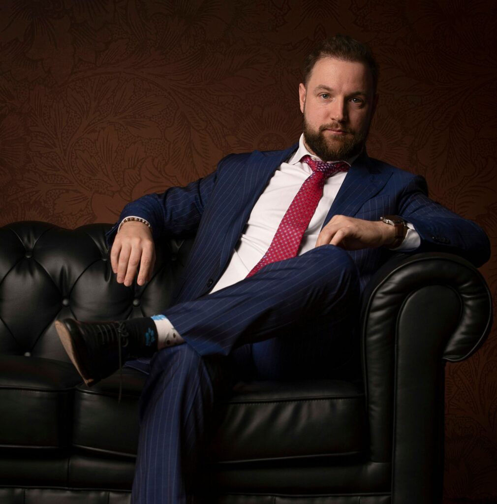 Un homme à la barbe et aux cheveux noirs, vêtu d'un costume bleu et d'une cravate rouge, est assis sur un canapé en cuir noir sur fond de motifs sombres. Il incarne l'éthique du bureau de style en rêvant à la création d'une marque de mode.