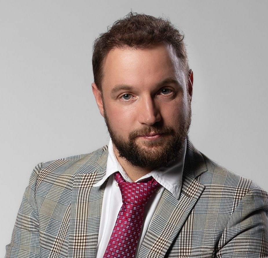 Un homme à la barbe et à la moustache courtes, portant une veste à carreaux, une chemise blanche et une cravate à motifs rouges, pose devant un fond gris uni.