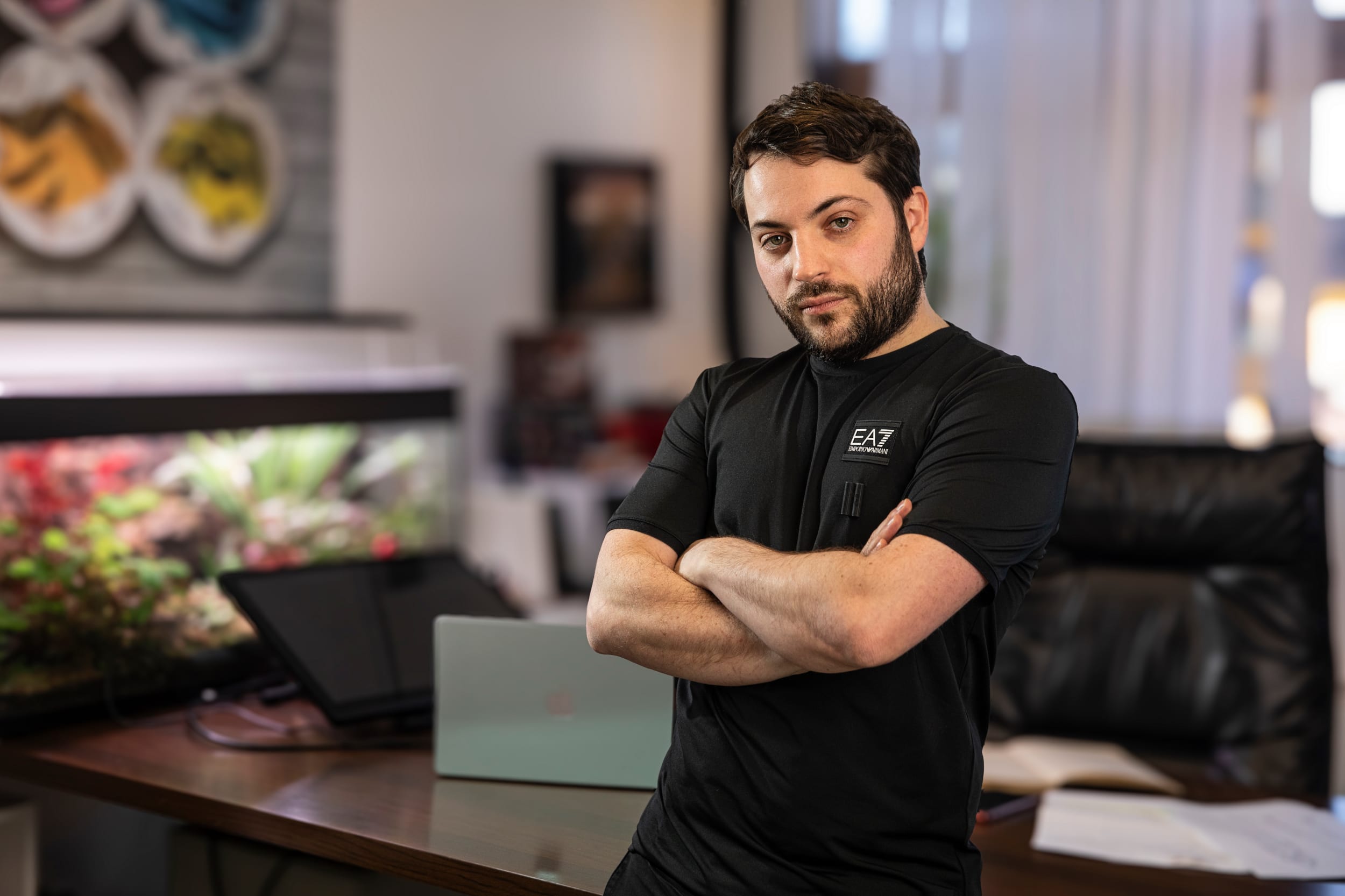 Un homme barbu se tient debout, les bras croisés, dans un bureau, à côté d'un ordinateur portable, d'un cahier ouvert et d'un aquarium en arrière-plan.