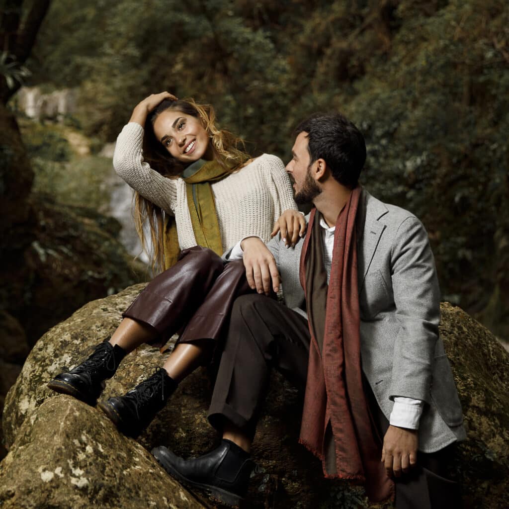 Une femme et un homme sont assis ensemble sur un gros rocher dans une zone boisée, tous deux vêtus de vêtements d'automne avec des écharpes et des foulards, entourés de verdure.