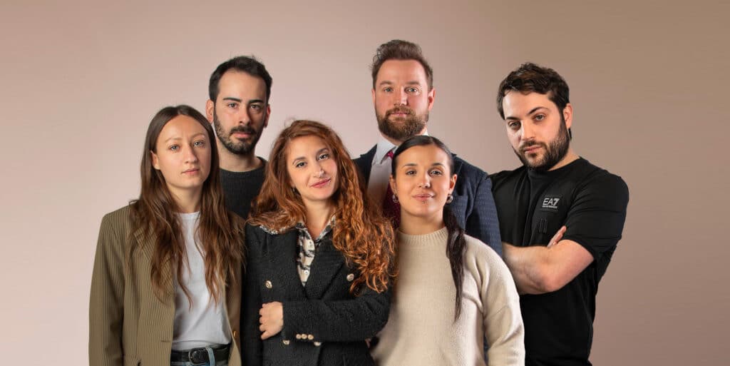 Six adults are standing close to each other against a plain background, facing the camera with neutral expressions.
