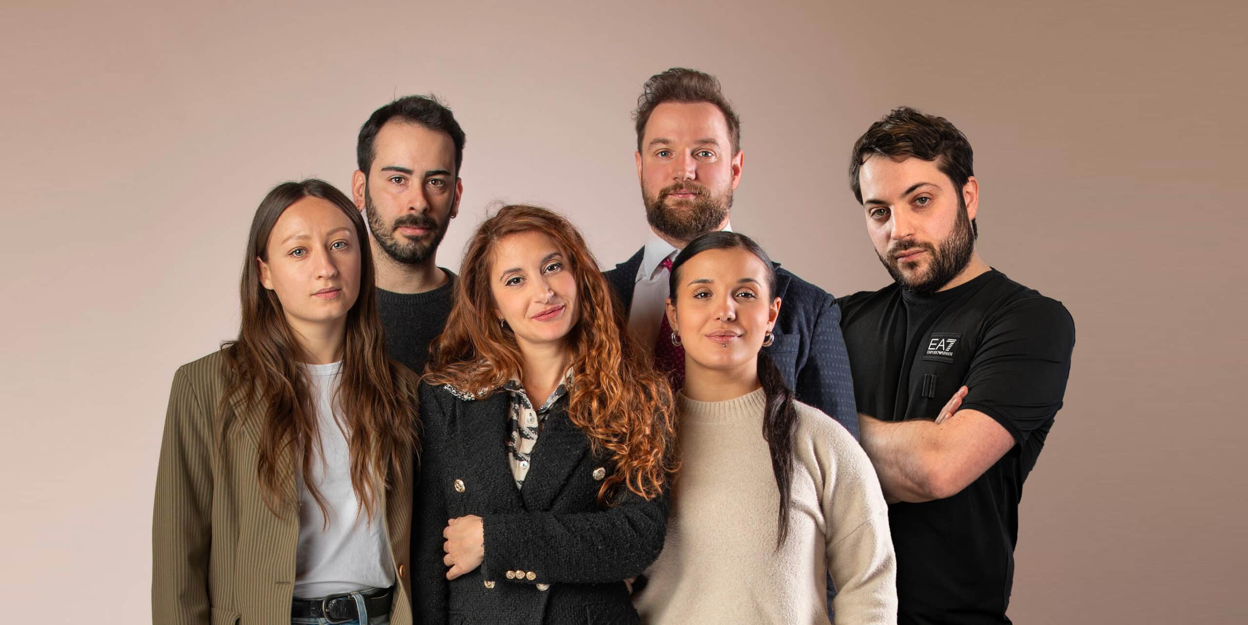 Six adultes se tiennent les uns à côté des autres sur un fond uni, faisant face à l'appareil photo avec des expressions neutres.