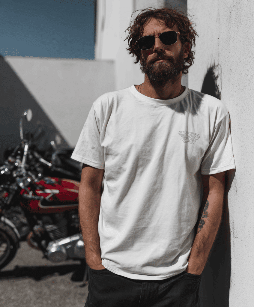 A man with a beard and sunglasses leans against a white wall with his hands in his pockets, wearing an Italian-made T-shirt; in the background are two motorbikes parked nearby.