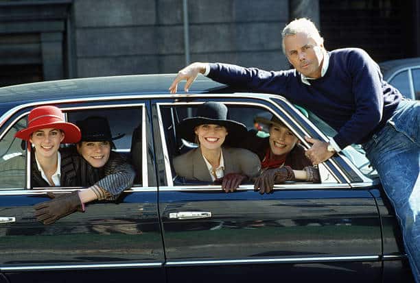 Four women in hats and gloves sit inside a car, smiling warmly as a man leans against the window: each exudes a timeless elegance reminiscent of a classic Armani moment.