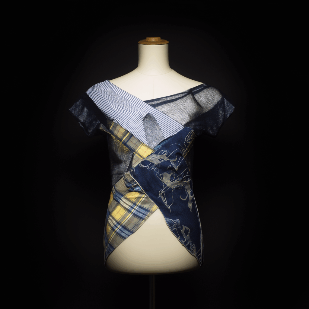 Un maniquí muestra un top asimétrico de manga corta confeccionado con tejidos mezclados como cuadros escoceses, vaqueros, rayas y estampados gráficos, una llamativa pieza que encarna la moda del upcycling sobre un fondo negro.