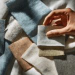 Eine Hand hält ein kleines Muster aus hellblauem Stoff über einer Reihe von Mustern verschiedener Webarten in neutralen und pastellfarbenen Tönen, die die kreativen Möglichkeiten des Upcycling von Mode zeigen.