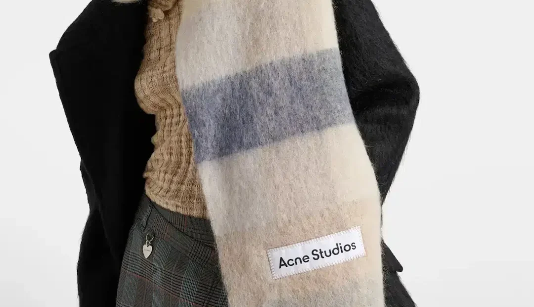 Eine Person, die einen schwarzen Mantel, einen beigen Pullover, einen karierten Rock und einen großen mehrfarbigen Acne Studios-Schal mit Fransen und einem sichtbaren Markenetikett trägt.